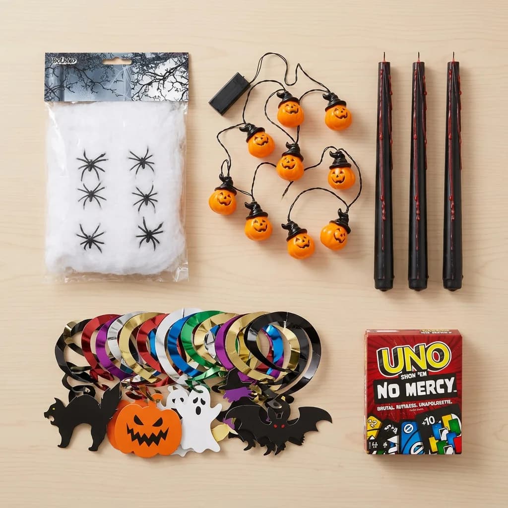 Halloween Party Deko: Spinnengewebe mit schwarzen Spinnen, Lichterkette mit Kürbissen, drei schwarze Kerzen, bunte Spiralgirlanden und UNO No Mercy Kartenspiel.