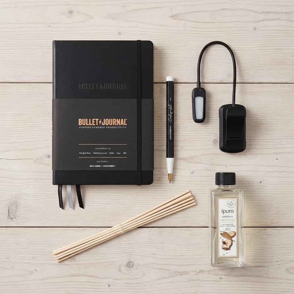 Fokus Box: schwarzes Bullet Journal A5, Gelschreiber, Klemmleuchte, Duftstäbchen-Flasche und Holzstäbchen auf hellem Holzuntergrund.