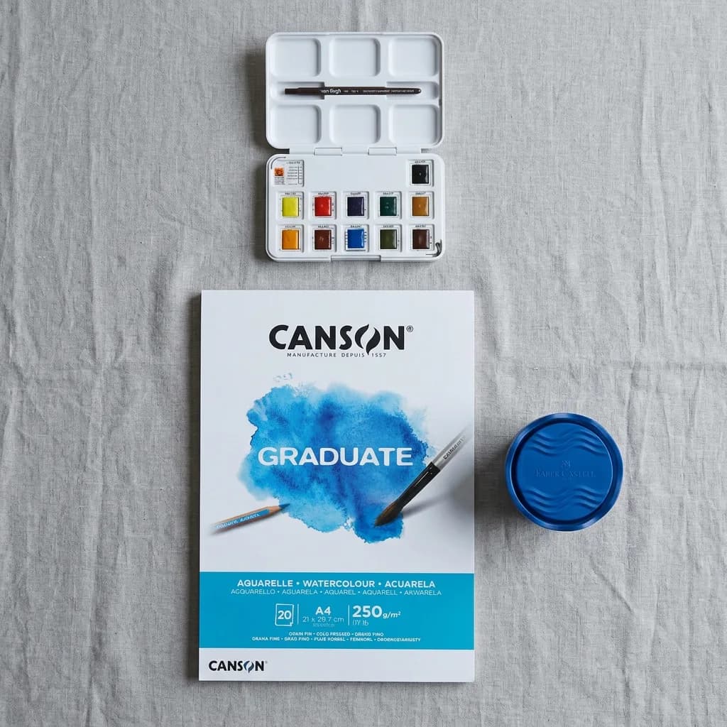 Aquarell malen: Aquarellfarb-Set mit 12 Näpfchen, Aquarellblock A4 20 Blatt und blauer Wasserbecher auf grauem Untergrund.