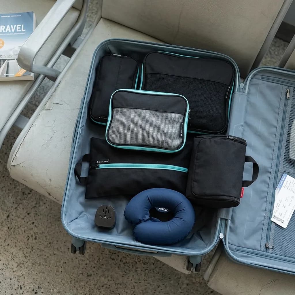 Handgepäck-Profi: Offener Koffer mit schwarzen Packwürfeln, Necessaire, Nackenkissen und Reiseadapter auf Flughafensitz.