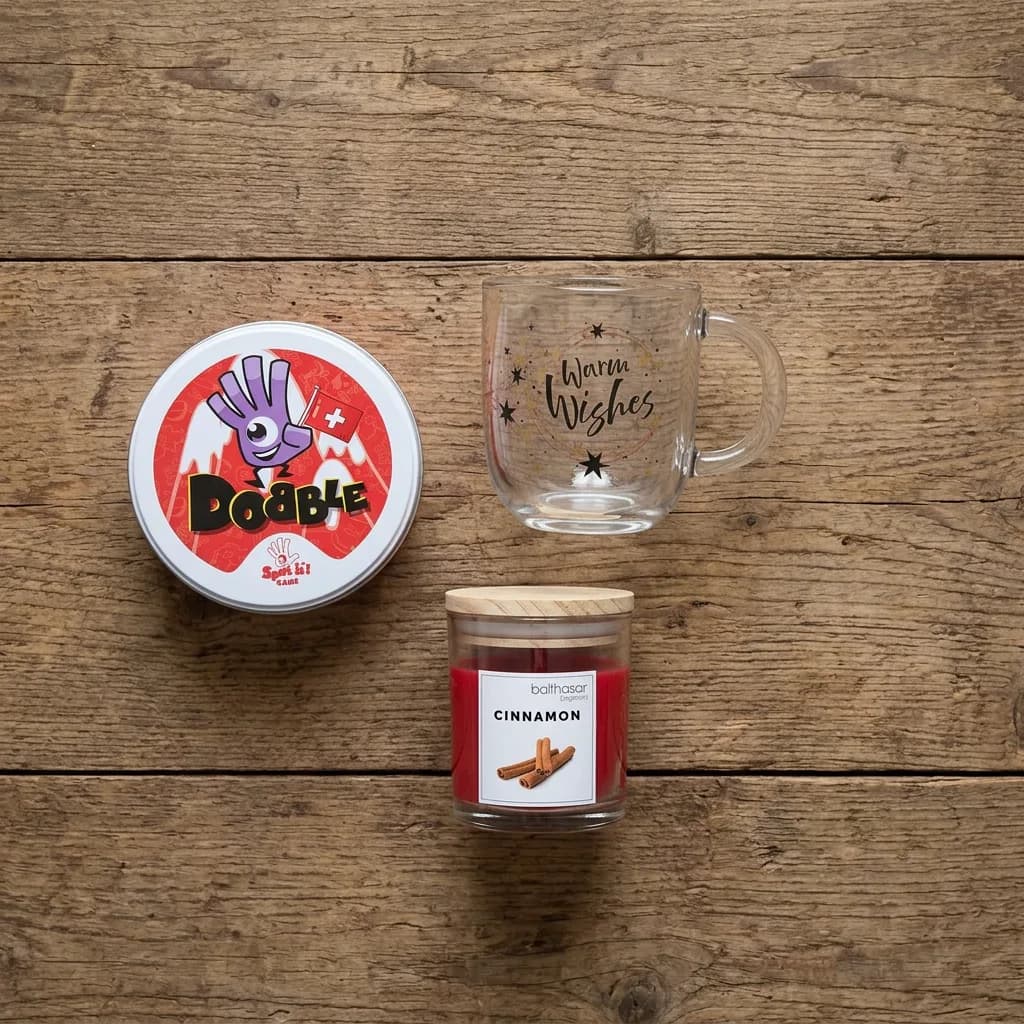 Wichtel-Geschenk unter 30: Kartenspiel Dobble Swiss in runder Metalldose, Glastasse «Warm Wishes» und rote Duftkerze Cinnamon mit Holzdeckel.