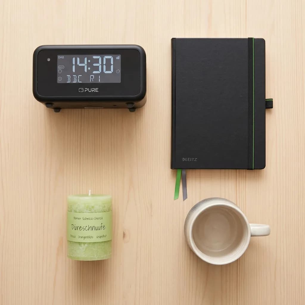 Morgenroutine ohne Handy: Radiowecker, schwarzes Notizbuch, hellgrüne Duftkerze und Kaffeetasse auf hellem Holzuntergrund.